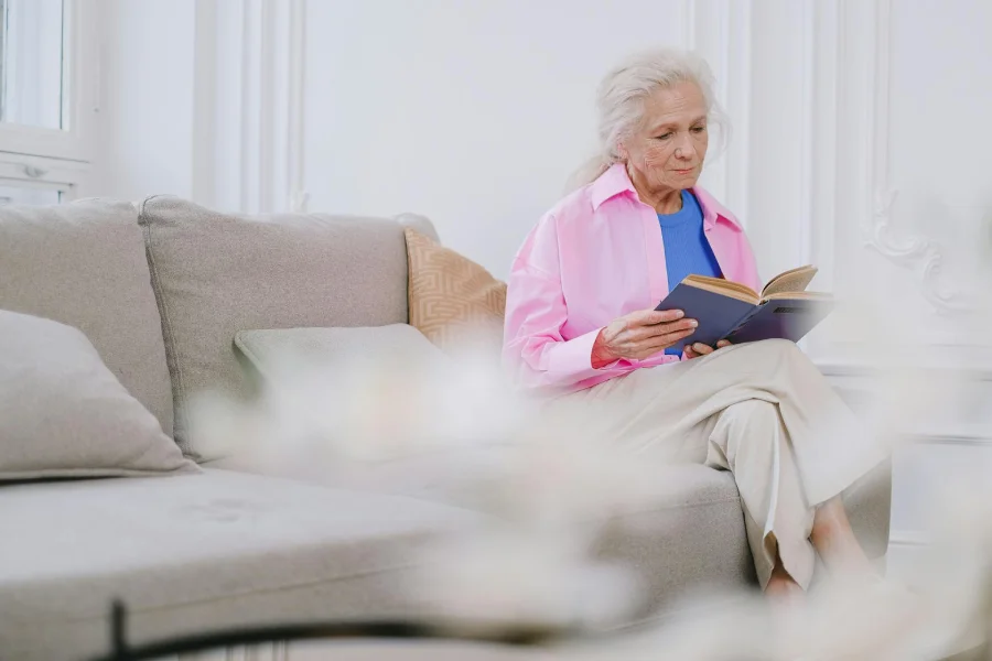 Elderly woman reading a book on a sofa in a bright living room.