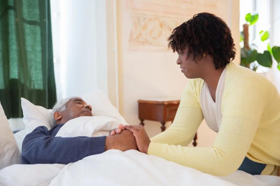 Caregiver holds an elderly patient’s hand beside a hospital bed.
