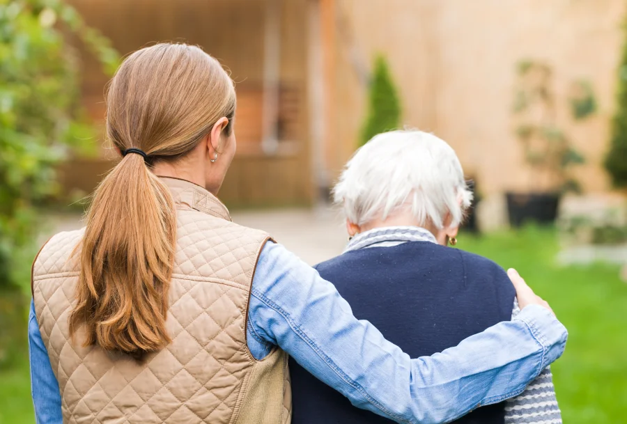 Caregiver walks with an elderly person outdoors, offering support.