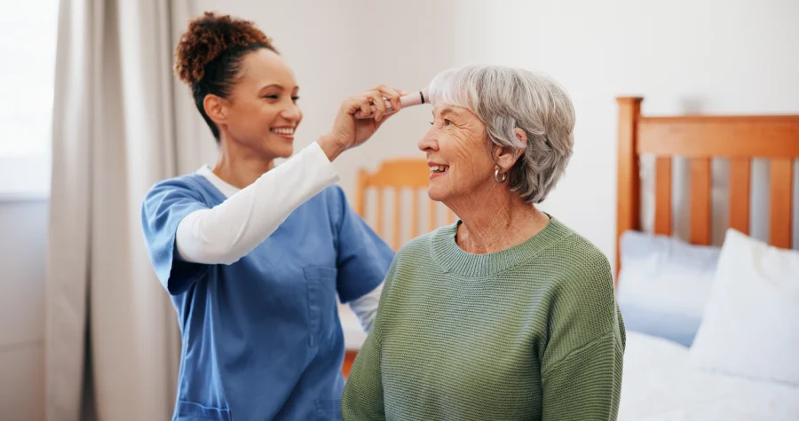 Caregiver smiling while checking an elderly woman’s forehead in a bedroom.