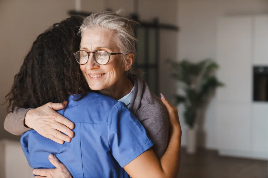 Smiling elderly woman hugs a caregiver in a bright room.